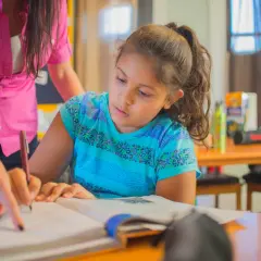 Dziewczynka z pomocą nauczycielki pisze w zeszycie. To przykład praktycznych zajęć podczas terapii pedagogicznej, studia podyplomowe.