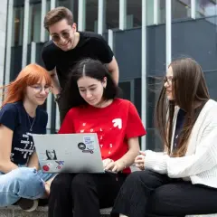 Cztery osoby siedzą na schodach przed budynkiem, patrząc na laptopa.