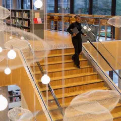 Kobieta schodzi po drewnianych schodach z laptopem. Zastanawia się, ile trwają studia podyplomowe, gdy mijają ją wiszące dekoracje w kształcie chmur.