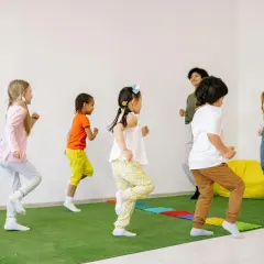 Dzieci tańczą na zielonej macie, a pomoc nauczyciela w przedszkolu czuwa nad zabawą.