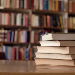 Stos książek na biurku, tło to regały pełne literatury. Idealne miejsce do nauki, gdy studiujesz turystykę.