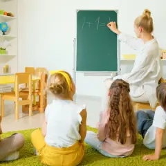 Nauczyciel wychowania przedszkolnego pisze na tablicy "1+1=2". Dzieci siedzą na dywanie i uważnie słuchają.
