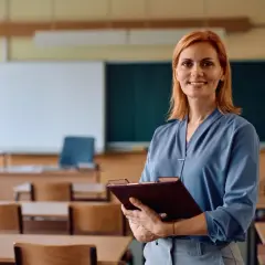 Uśmiechnięta nauczycielka z rudymi włosami trzyma teczkę w pustej sali lekcyjnej. Czas na zasłużony urlop nauczyciela!