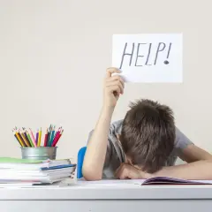 Chłopiec z kartką "HELP!" nad głową, przy biurku pełnym książek i kredek. Może to być sygnał trudności, wymagający diagnozy psychologicznej.