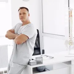 Mężczyzna w uniformie medycznym opiera się o biurko, na którym stoi model kręgosłupa. To przyszły specjalista, który wybrał fizjoterapia studia.