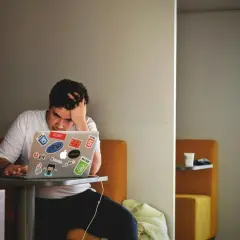 Młody programista z ręką we włosach, wyraźnie odczuwający **stres w pracy**. Na laptopie mnóstwo naklejek z technologiami.