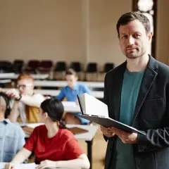 Nauczyciel z książką w ręku, przygotowujący się do lekcji. W tle widać uczniów, co sugeruje jego pensum nauczyciela.