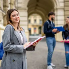 Uśmiechnięta studentka z notatnikiem w ręku, przygotowująca się do studiów 2 stopnia. W tle para studentów.
