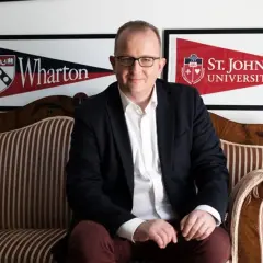 Mężczyzna w garniturze siedzi na kanapie. W tle banery Wharton i St. John's University, prestiżowa uczelnia w USA.