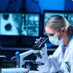 Diagnosta laboratoryjny w okularach i masce analizuje próbki pod mikroskopem. Na ekranach komputera widać wyniki badań.