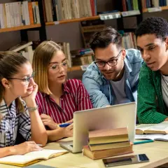 Studenci na studiach humanistycznych pochylają się nad laptopem w bibliotece, dyskutując o projekcie.