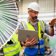 Doświadczony monter w kamizelce odblaskowej i kasku, z laptopem w ręku, obsługuje maszynę. Obok wachlarz banknotów 100 zł.