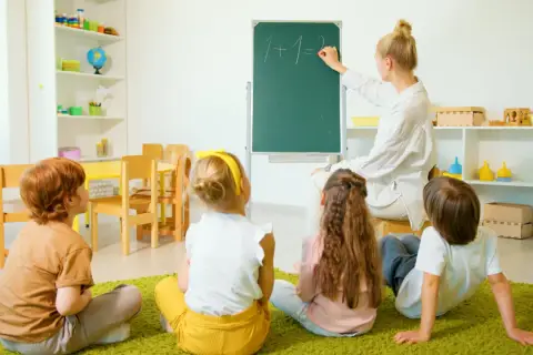 Nauczyciel wychowania przedszkolnego pisze na tablicy "1+1=2". Dzieci siedzą na dywanie i uważnie słuchają.