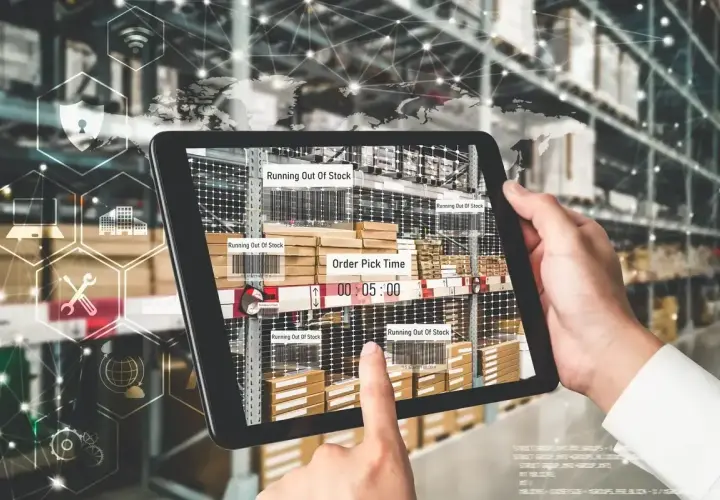 Ręce trzymają tablet z widokiem magazynu, pokazujący alerty "Running Out Of Stock" i czas kompletacji zamówienia. To przykład nowoczesnego zarządzania łańcuchem dostaw.