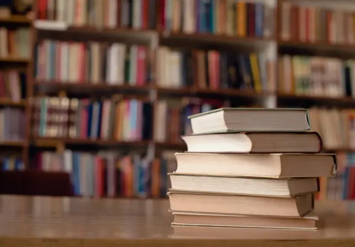 Stos książek na biurku, tło to regały pełne literatury. Idealne miejsce do nauki, gdy studiujesz turystykę.