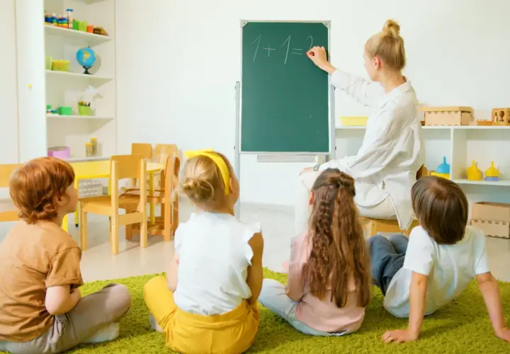 Nauczyciel wychowania przedszkolnego pisze na tablicy "1+1=2". Dzieci siedzą na dywanie i uważnie słuchają.