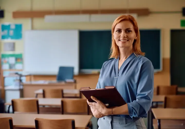 Uśmiechnięta nauczycielka z rudymi włosami trzyma teczkę w pustej sali lekcyjnej. Czas na zasłużony urlop nauczyciela!