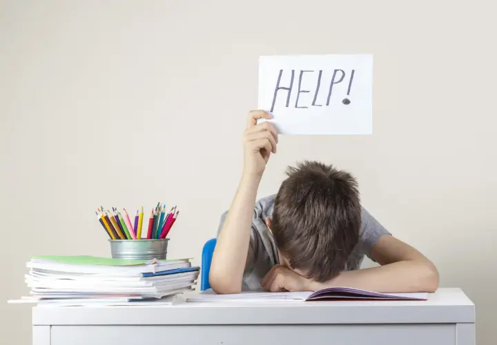 Chłopiec z kartką "HELP!" nad głową, przy biurku pełnym książek i kredek. Może to być sygnał trudności, wymagający diagnozy psychologicznej.