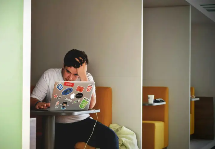 Młody programista z ręką we włosach, wyraźnie odczuwający **stres w pracy**. Na laptopie mnóstwo naklejek z technologiami.