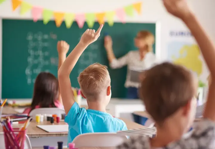 Uczniowie z podniesionymi rękami słuchają uważnie, co mówi wychowawca przy tablicy.