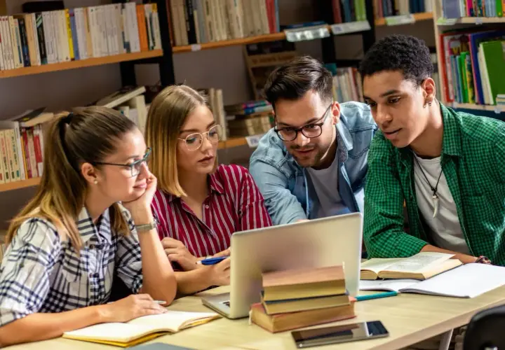 Studenci na studiach humanistycznych pochylają się nad laptopem w bibliotece, dyskutując o projekcie.