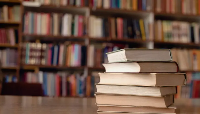 Stos książek na biurku, tło to regały pełne literatury. Idealne miejsce do nauki, gdy studiujesz turystykę.