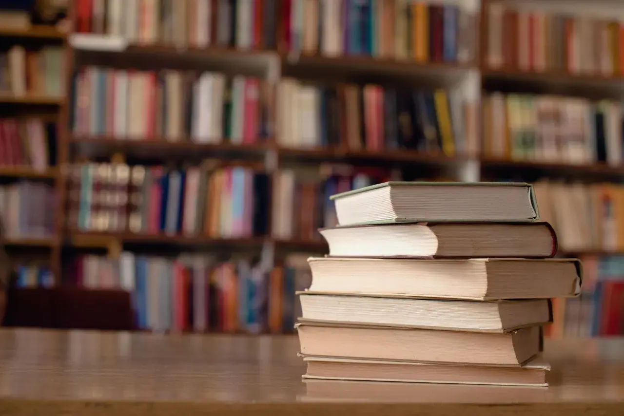 Stos książek na biurku, tło to regały pełne literatury. Idealne miejsce do nauki, gdy studiujesz turystykę.