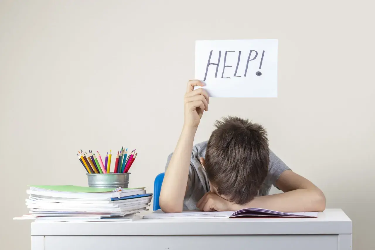 Chłopiec z kartką "HELP!" nad głową, przy biurku pełnym książek i kredek. Może to być sygnał trudności, wymagający diagnozy psychologicznej.