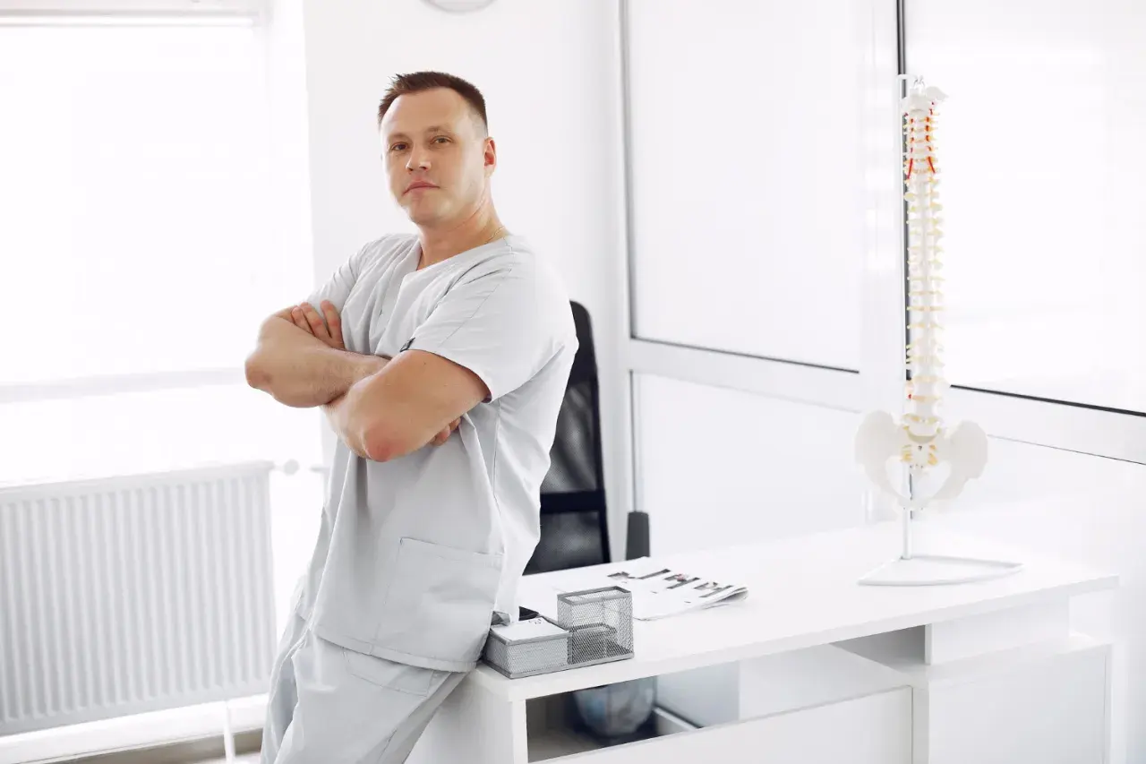 Mężczyzna w uniformie medycznym opiera się o biurko, na którym stoi model kręgosłupa. To przyszły specjalista, który wybrał fizjoterapia studia.