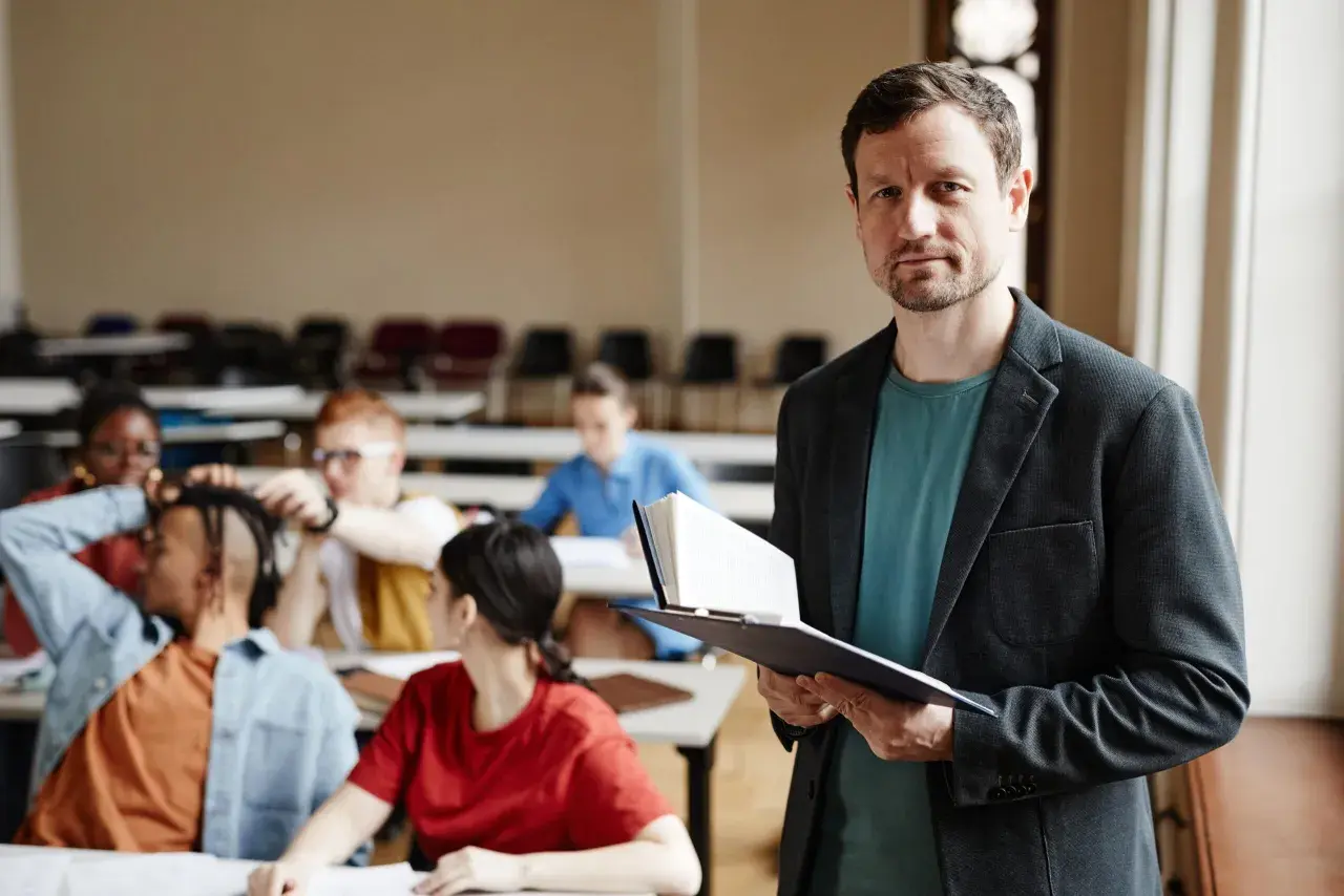 Nauczyciel z książką w ręku, przygotowujący się do lekcji. W tle widać uczniów, co sugeruje jego pensum nauczyciela.