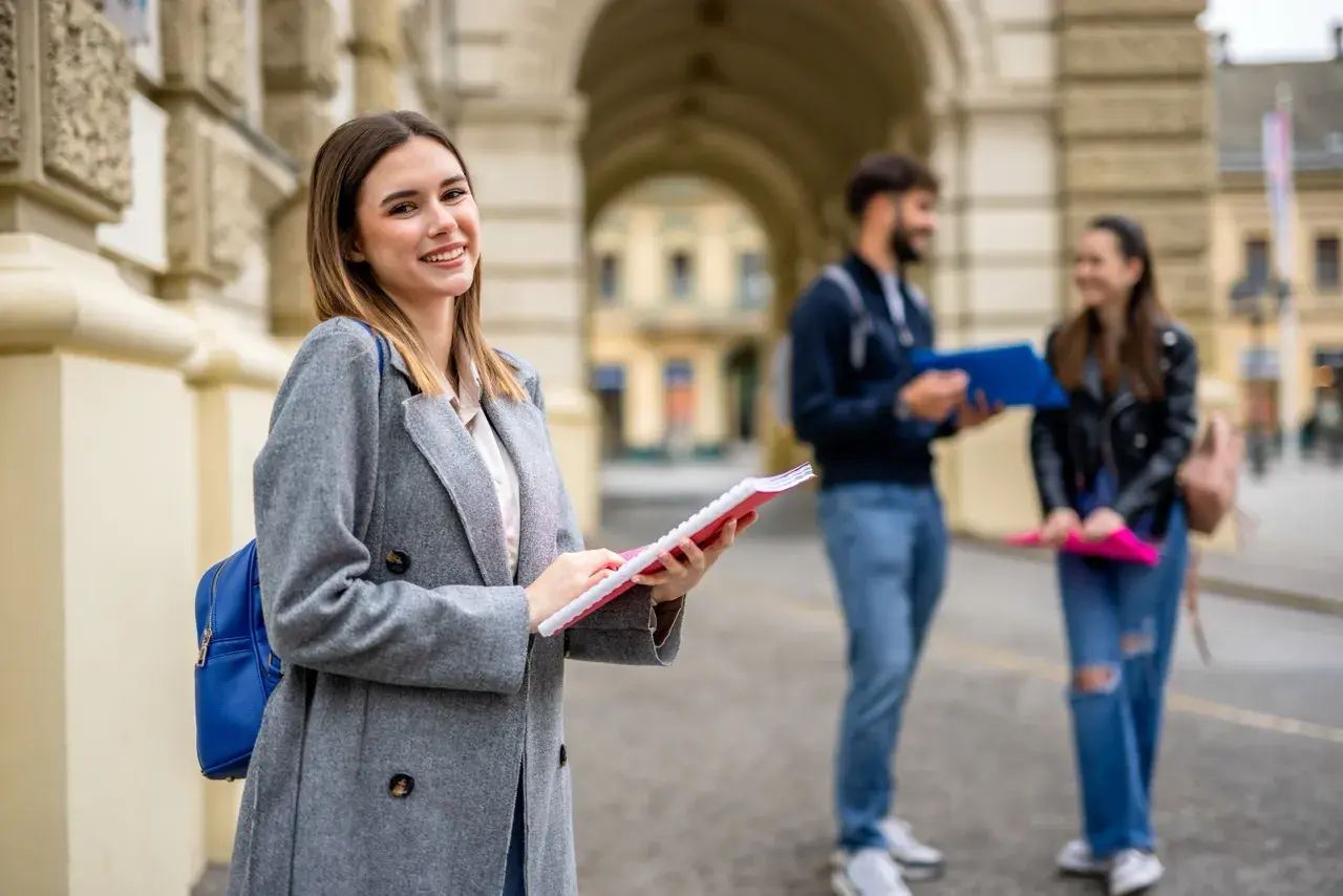 Uśmiechnięta studentka z notatnikiem w ręku, przygotowująca się do studiów 2 stopnia. W tle para studentów.