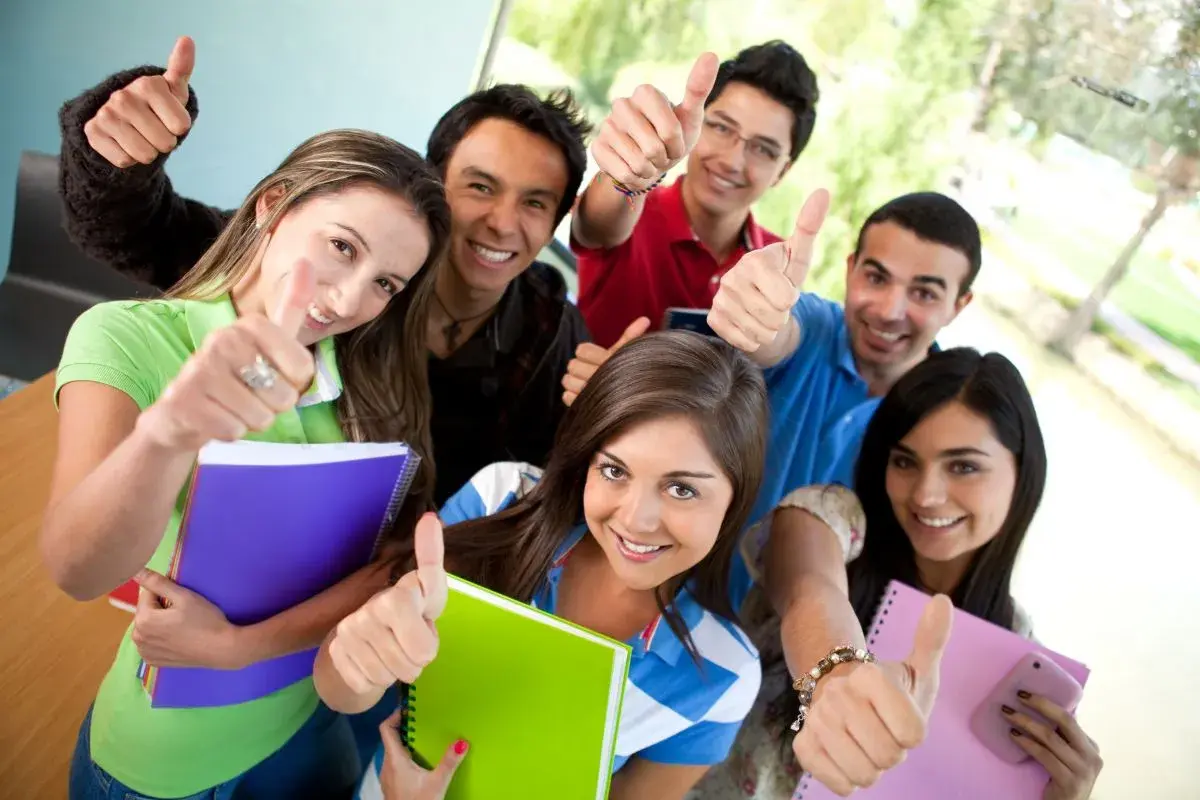 Uśmiechnięci studenci z notatnikami pokazują kciuki w górę, gotowi na studia stacjonarne.