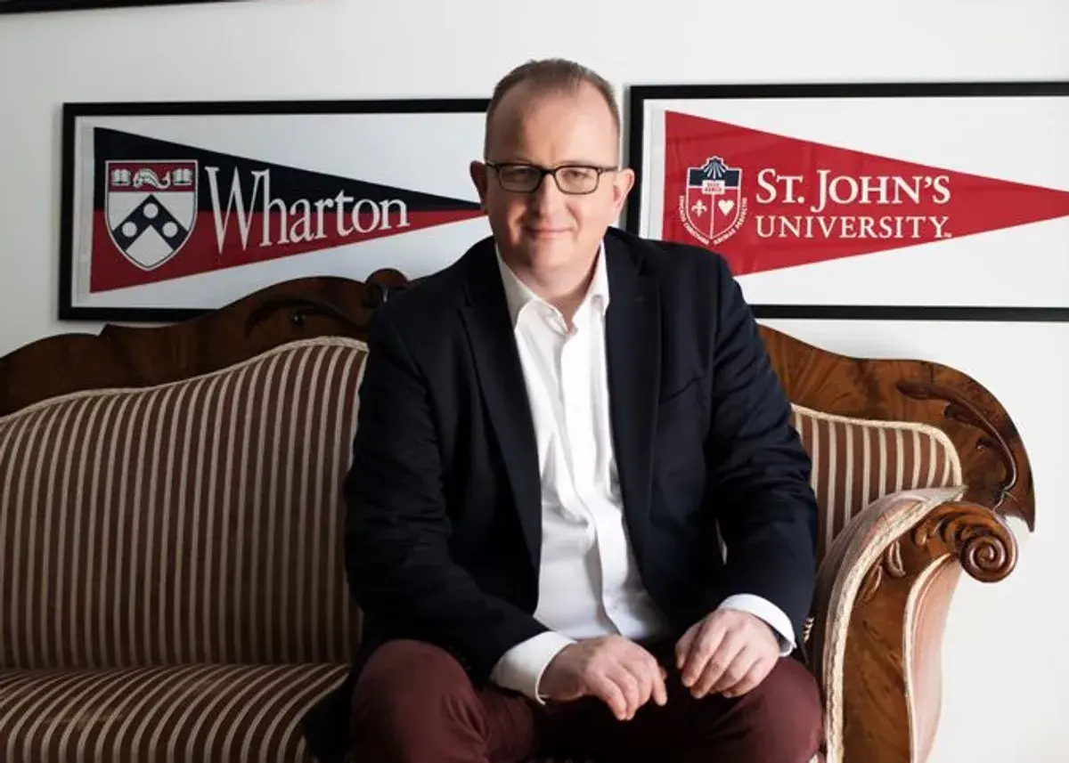 Mężczyzna w garniturze siedzi na kanapie. W tle banery Wharton i St. John's University, prestiżowa uczelnia w USA.