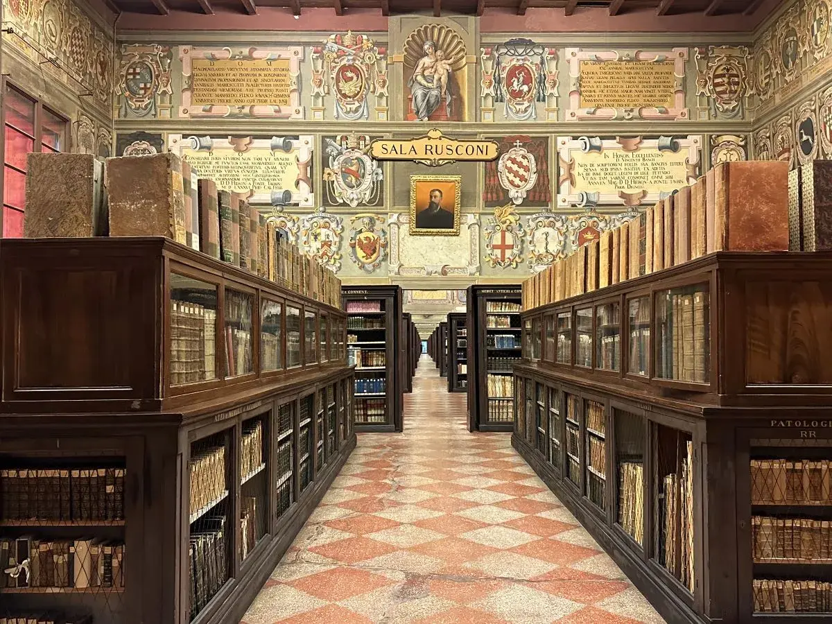 Wnętrze zabytkowej biblioteki z rzędami drewnianych regałów pełnych starych ksiąg, ozdobione malowidłami. To miejsce, gdzie narodziła się nauka, symbolizujące najstarszy uniwersytet na świecie.