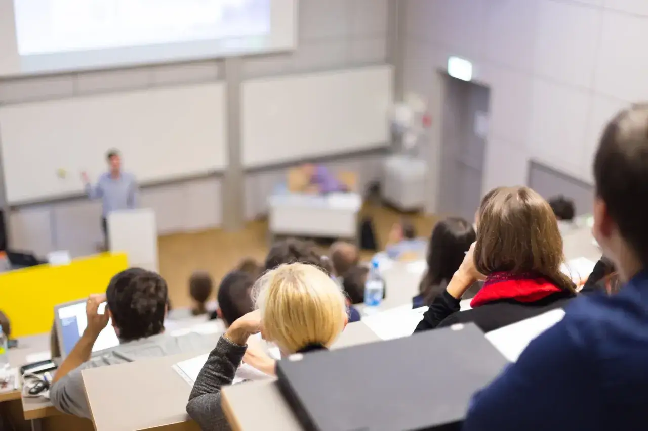 Studenci na wykładzie podczas studiów stacjonarnych. Wykładowca stoi przy tablicy, a słuchacze notują.