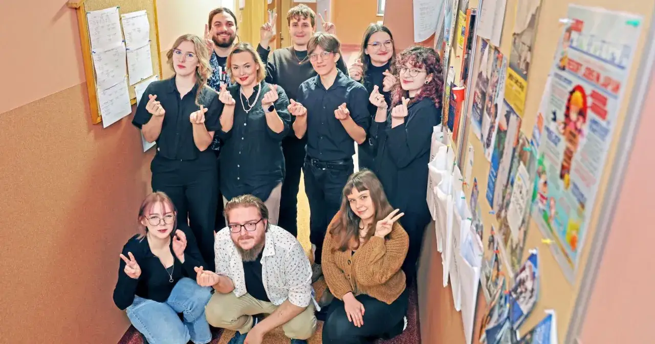 Grupa studentów japonistyki pozuje do zdjęcia, pokazując gesty serca. Na ścianie wiszą plakaty związane z Japonią.