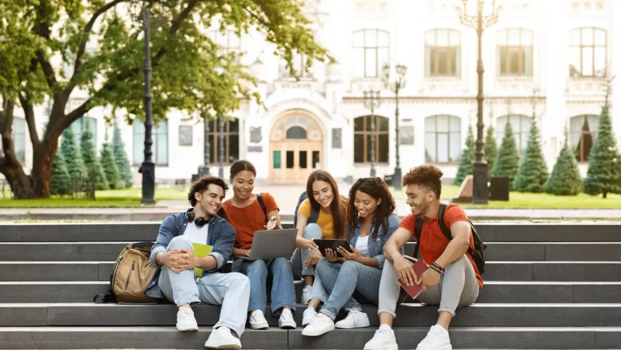 Studenci na schodach przed budynkiem prestiżowej uczelni w USA. Wspólnie pracują przy laptopie i tablecie, ciesząc się słońcem.