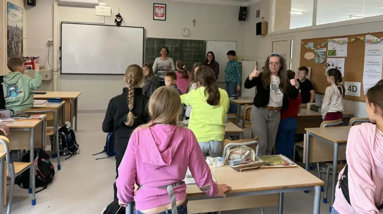 Nauczycielka z uśmiechem i kciukami w górze prowadzi lekcję, a uczniowie aktywnie uczestniczą w zajęciach. To obrazek z życia, gdzie pedagogika studia nabiera praktycznego wymiaru.