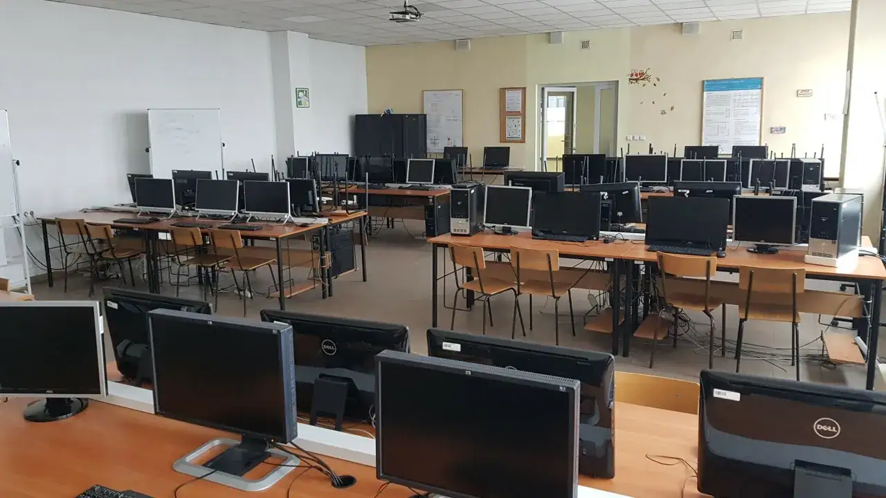 Pusta sala komputerowa, idealna do studiów programistycznych. Wiele stanowisk z monitorami i krzesłami czeka na studentów.