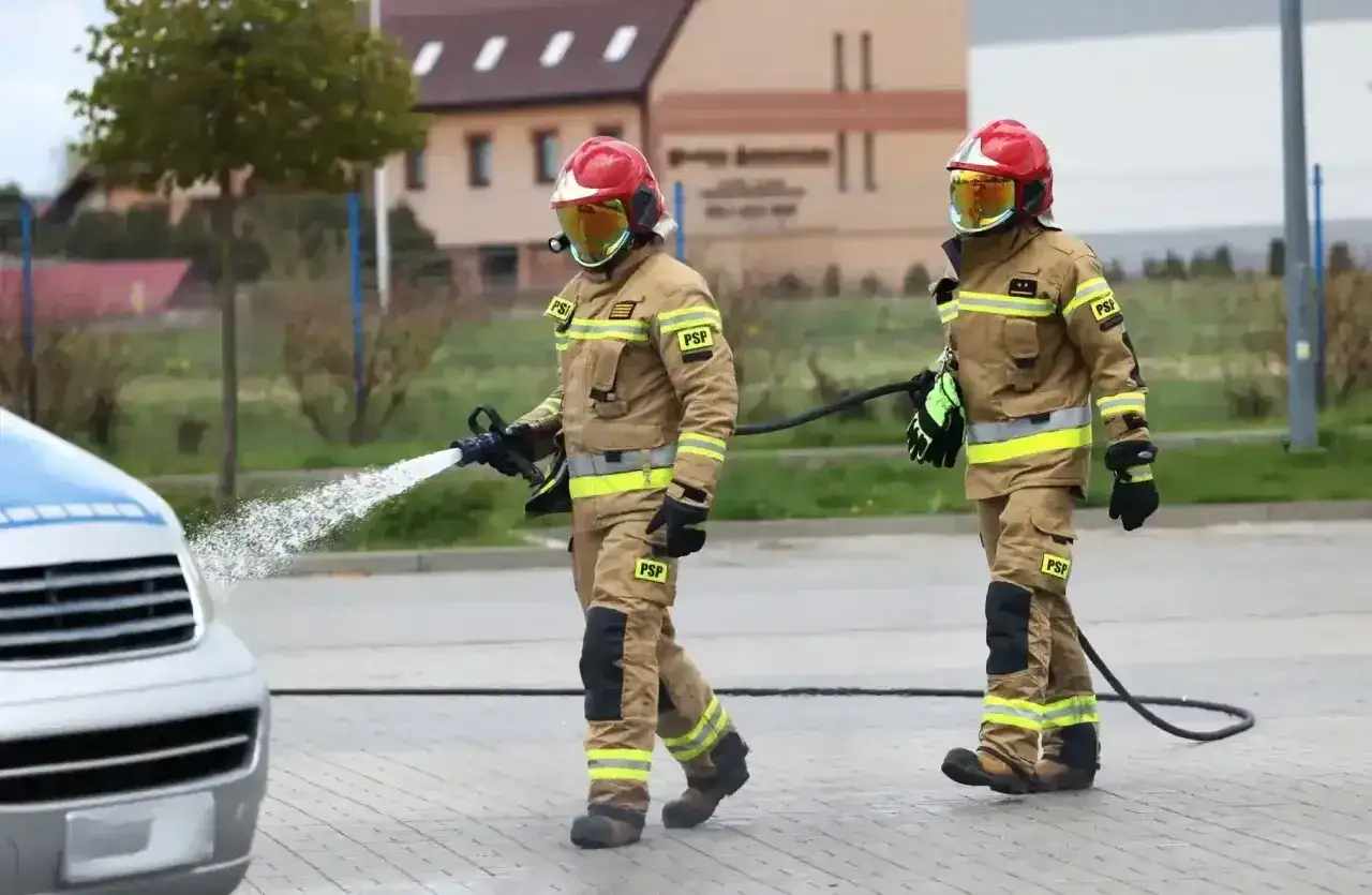 Strażacy w pełnym rynsztunku ćwiczą gaszenie pożaru. Jeden z nich kieruje strumień wody na pojazd, drugi niesie wąż. To część ich studiów strażackich.