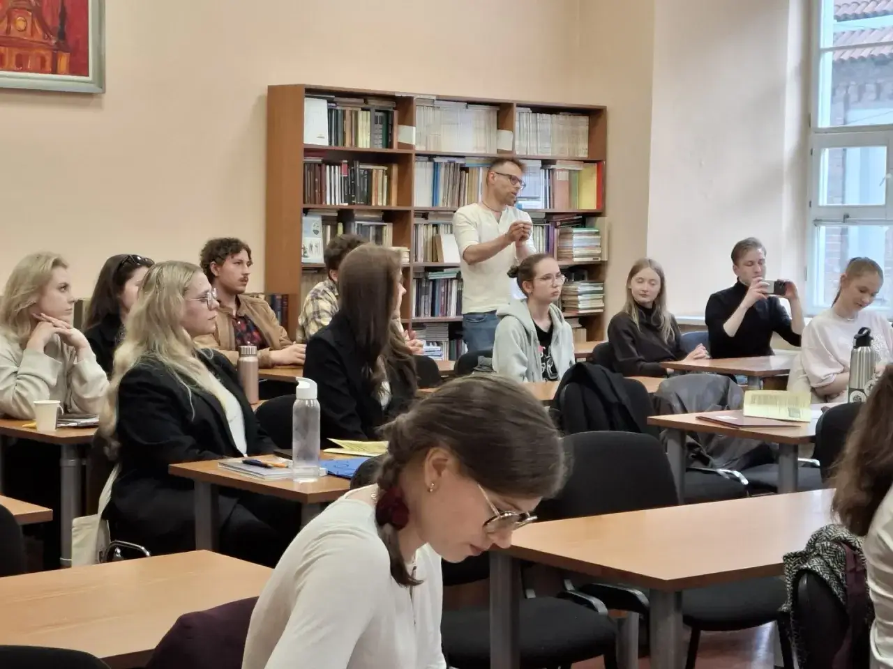 Wykład z filologii polskiej. Studenci słuchają wykładowcy przy regałach pełnych książek.