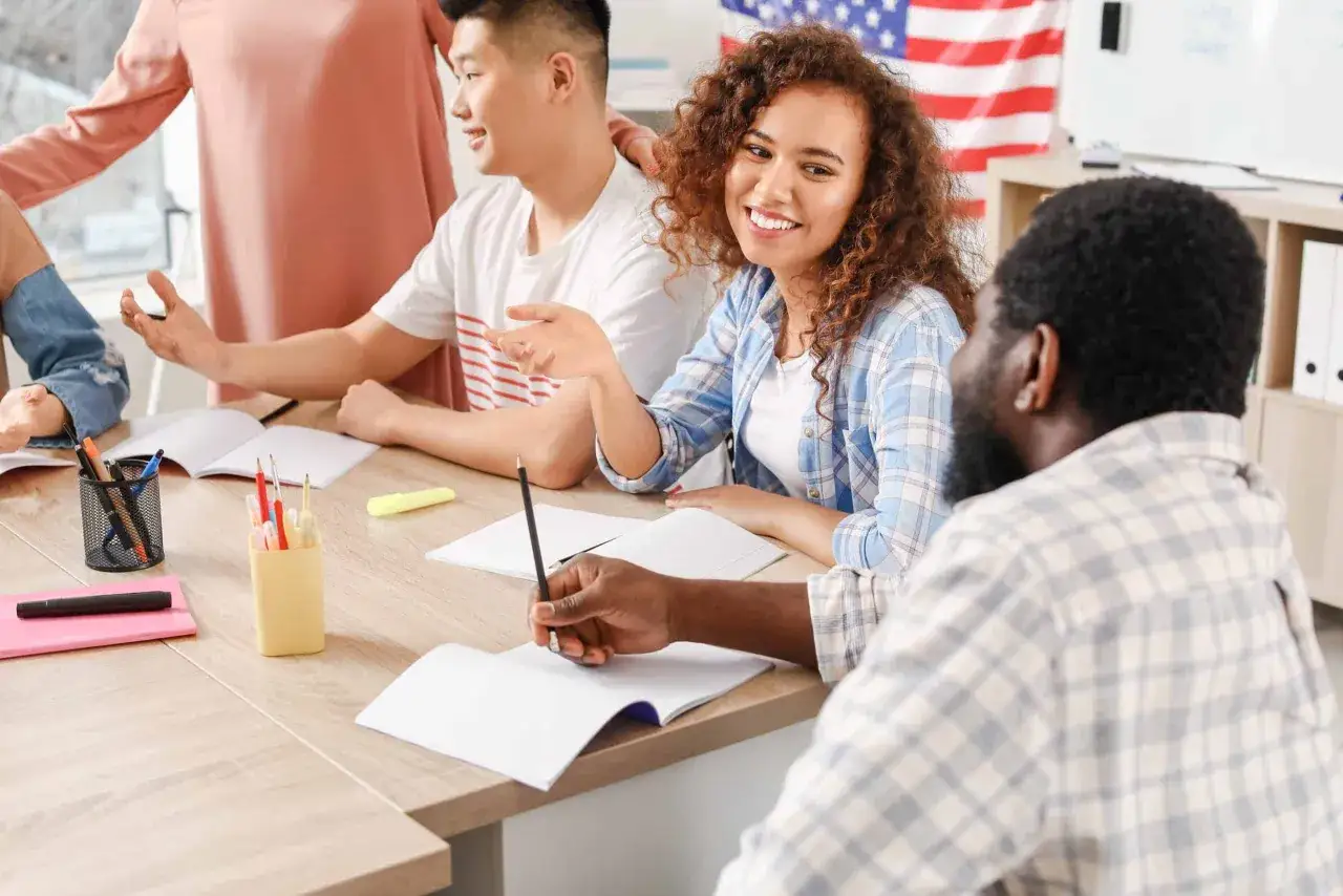 Uśmiechnięta studentka z kręconymi włosami podczas zajęć. Grupa osób przy stole z notatkami i flagą USA w tle. To idealne miejsce na studia niestacjonarne.