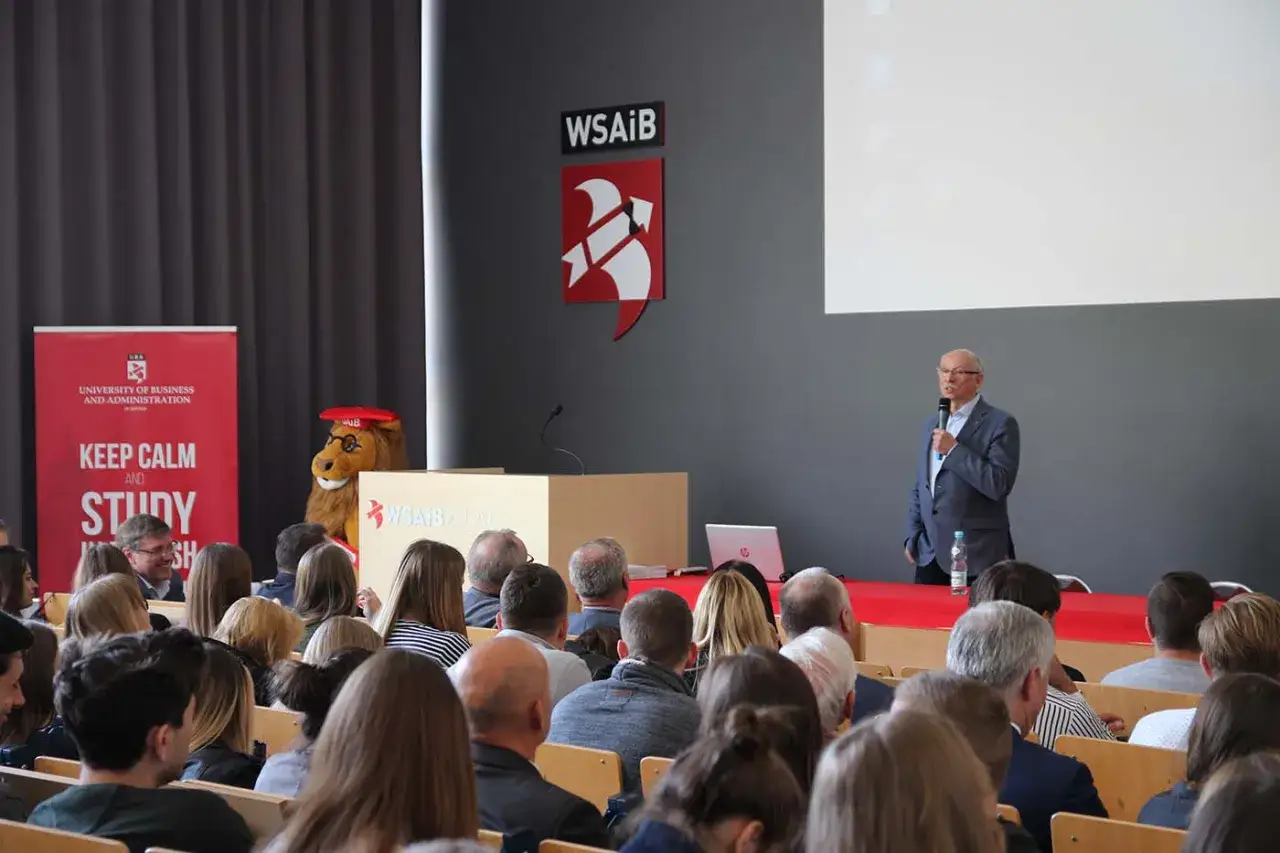 Wykład na uczelni WSAiB. Studenci administracji słuchają prelegenta. Maskotka lwa zachęca do nauki.