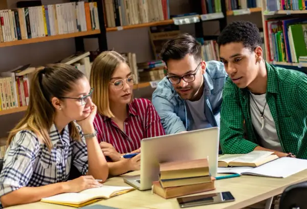 Studenci na studiach humanistycznych pochylają się nad laptopem w bibliotece, dyskutując o projekcie.